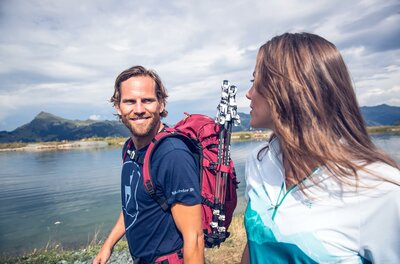 Zwei Wanderer genießen die Aussicht am Ufer eines Gewässers. Die Stimmung ist entspannt und freundlich.