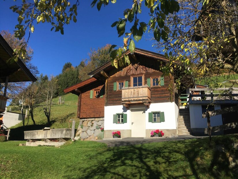 Alpines Holzhaus mit weißem Sockelgeschoss, grünen Fensterläden und Holzbalkon vor blauem Himmel.