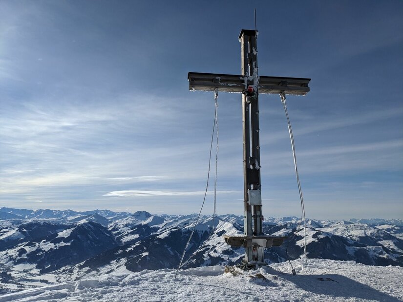 Ein hölzernes Gipfelkreuz auf einem schneebedeckten Berg vor einem Panorama aus verschneiten Alpengipfeln.