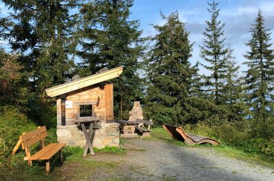 Kleine Holzhütte mit Sitzbänken und einer Holzliege an einem Schotterweg vor Nadelbäumen.