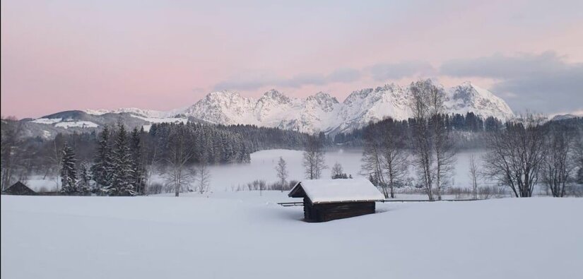 Winterlandschaft am Schwarzsee Verschneite Wiese mit kleiner Holzhütte vor bewaldeten Hügeln und schneebedecktem Gebirgsmassiv unter rosa Himmel.
