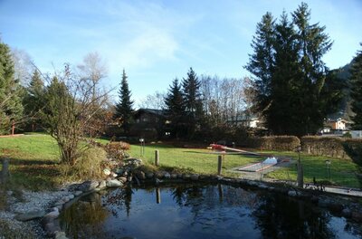 Minigolfpark Bruggerhof Ein kleiner Teich im Vordergrund mit einer Minigolfanlage auf einer Rasenfläche dahinter.