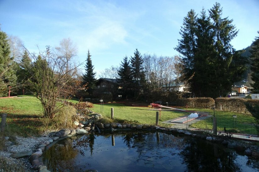 Minigolfpark Bruggerhof Ein kleiner Teich im Vordergrund mit einer Minigolfanlage auf einer Rasenfläche dahinter.