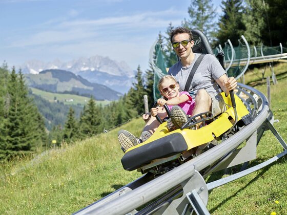 Timoks Coaster Sommer Eine Familie fährt mit einer Rodelbahn auf einem Berg. Im Hintergrund sind grüne Wiesen und hohe Berge zu sehen.