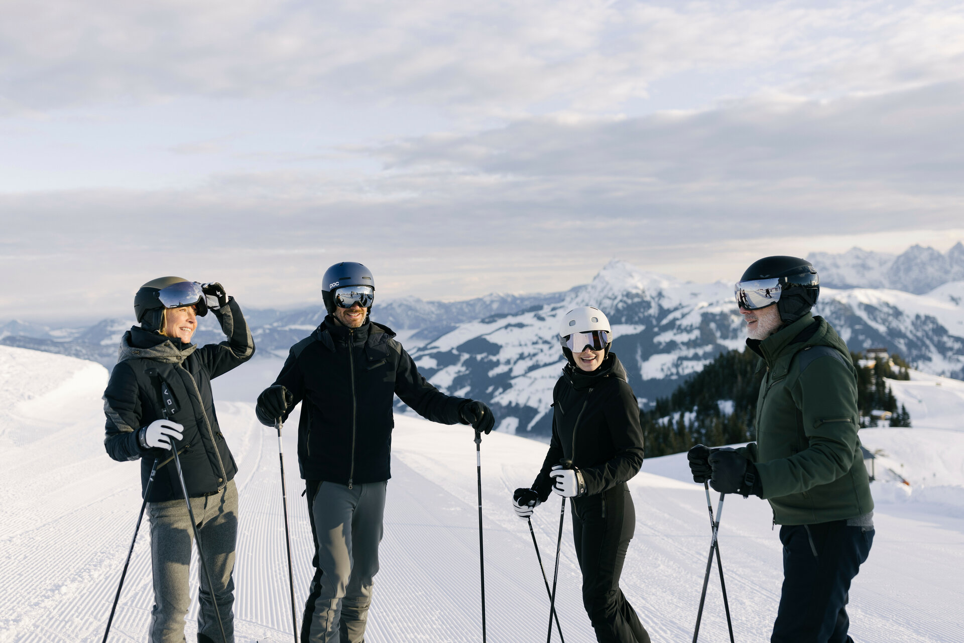 Eine Gruppe von vier Personen in Skiausrüstung lacht und unterhält sich auf einem sonnigen Berggipfel.