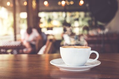 <p>Eine Tasse Kaffee auf einem Tisch mit Holzoberfläche. Im Hintergrund sind Personen in einem Café zu sehen.</p>