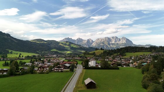Blick auf ein Dorf im Tal mit grünen Wiesen und dem Gebirgsmassiv des Wilden Kaisers im Hintergrund.