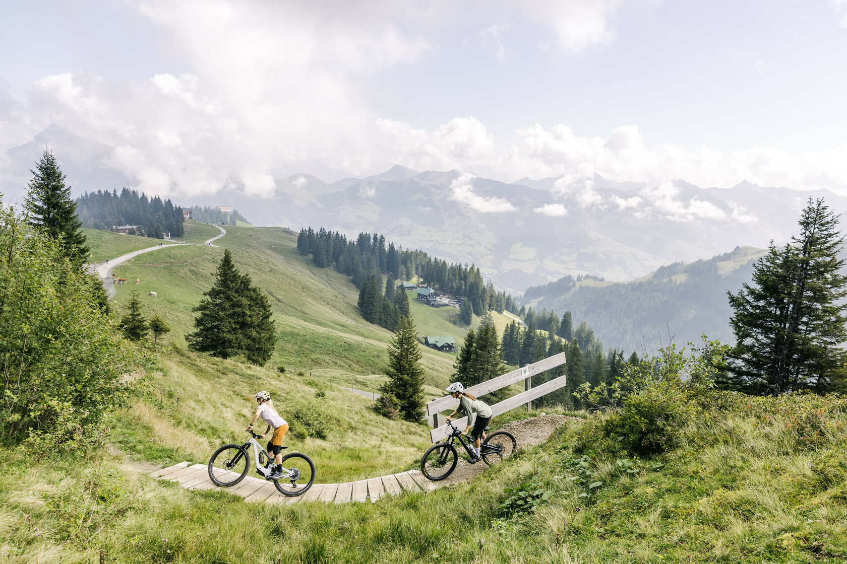 Zwei Mountainbiker fahren auf dem Hahnenkamm Trail in Kitzbühel über einen kurvenreichen Holzsteg durch eine grüne Berglandschaft mit bewaldeten Hügeln.