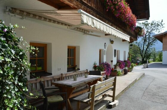 Terrasse am Malernhof Kitzbühel Holztisch und Bänke auf einer Terrasse unter einer Markise vor einem weißen Bauernhaus.