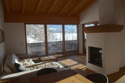 Wohnraum Apartment Hofer Manfred Moderner Wohnraum mit Sofa, Kamin und großem Panoramafenster mit Blick auf eine verschneite Winterlandschaft.