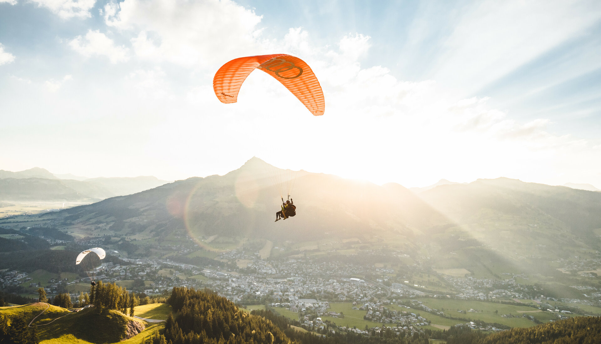 Ein Tandem-Paraglider fliegt über ein grünes Tal in Kitzbühel, umgeben von Bergen bei Sonnenschein.