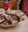Eine Hand serviert mit einem Pfannenwender Kaiserschmarrn aus einer Metallpfanne auf einem gedeckten Tisch.
