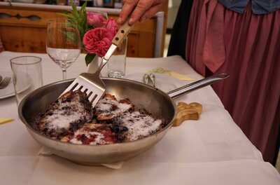 Eine Hand serviert mit einem Pfannenwender Kaiserschmarrn aus einer Metallpfanne auf einem gedeckten Tisch.
