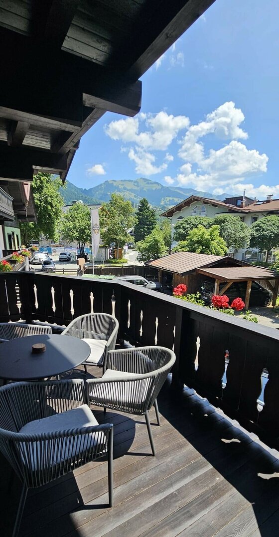 Holzbalkon mit modernen Sitzmöbeln und Blick auf Berge und umliegende Häuser unter blauem Himmel.