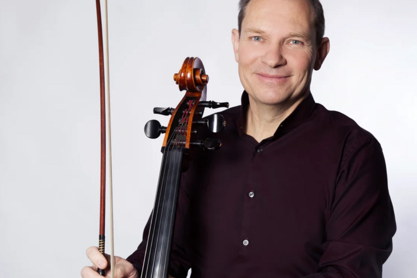 Ein Mann im dunklen Hemd hält ein Cello und einen Bogen vor weißem Hintergrund.