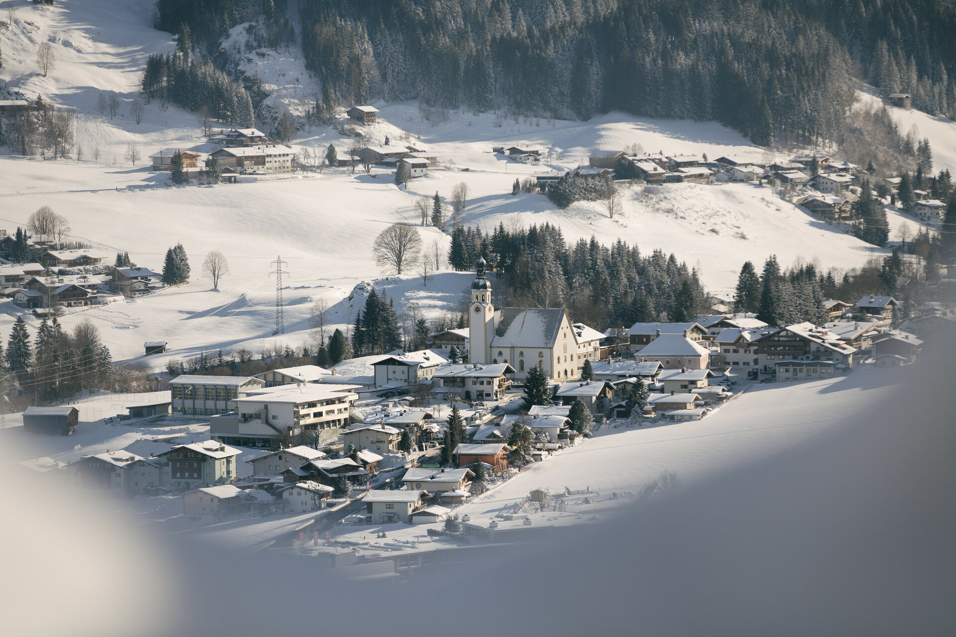 Winterlandschaft in Jochberg bei Kitzbühel mit Dorf, Bergen und Nadelbäumen.