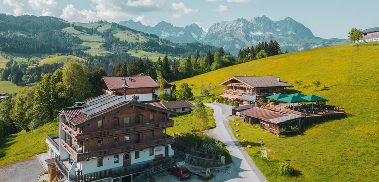 Zwei rustikale Holzhäuser stehen in einer idyllischen Umgebung, umgeben von sanften Hügeln und majestätischen Bergen.