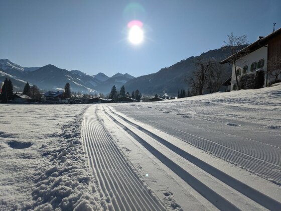 Stadtloipe Kitzbühel-Aurach-Jochberg Präparierte Langlaufloipe im Schnee mit Blick auf Berge und Häuser unter sonnigem Himmel.