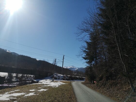 Wegverlauf bei der Labalm-Tour Asphaltierter Weg neben einer Bahnbrücke und verschneiten Wiesen unter blauem Himmel.