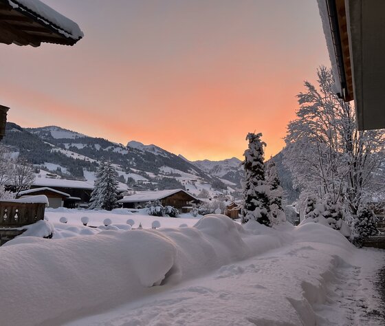 Verschneite Alpendorf-Landschaft mit Holzhäusern und Bergen vor einem orangefarbenen Abendhimmel.
