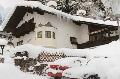 Schneebedecktes alpines Wohnhaus mit Eiszapfen am Dach und eingeschneiten Autos im Vordergrund.