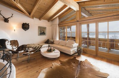 Innenansicht KitzChalet Reith Rustikaler Wohnraum mit weißem Sofa, Holzboden, Fellteppich und großen Glasfronten mit Blick auf eine Schneelandschaft.