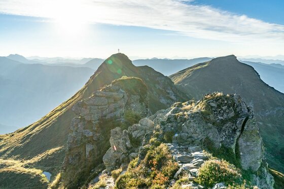 Gipfelpanorama der Kitzbüheler Südberge Felsiger Gebirgsgrat mit Gipfelkreuz im Gegenlicht vor weiteren Bergketten.