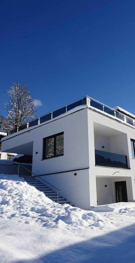 Apartment Sabrina im Winter Modernes weißes Apartmentgebäude mit Flachdach und Glasbalkonen im Schnee unter blauem Himmel.