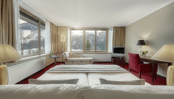 Zimmer im Schlosshotel Lebenberg Hotelzimmer mit Doppelbett, Schreibtisch und Blick durch große Fenster auf eine verschneite Berglandschaft.
