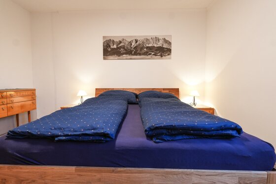 Ein gemütliches Schlafzimmer mit einem großen Doppelbett und blauen Decken. An den Wänden hängen Bilder, und das Licht ist sanft.