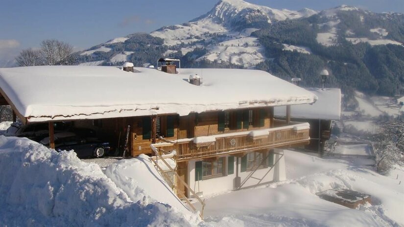 Schneebedecktes Holzchalet mit Balkon vor einer alpinen Bergkulisse im Winter.