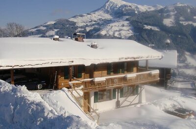 Schneebedecktes Holzchalet mit Balkon vor einer alpinen Bergkulisse im Winter.
