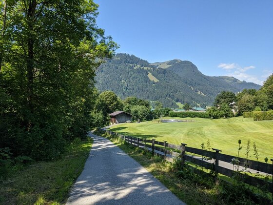 Wanderweg bei Aurach Asphaltierter Wanderweg neben einer grünen Wiese mit Holzzaun und bewaldeten Bergen im Hintergrund.