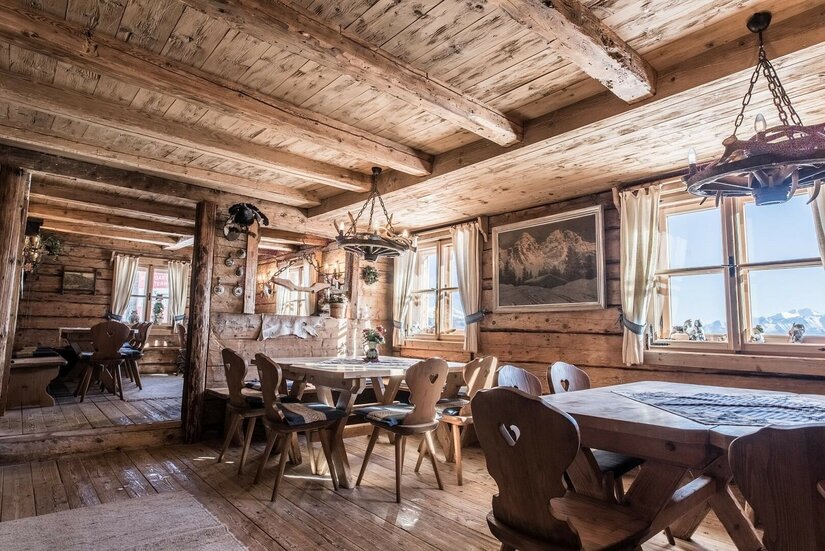 Rustikale Gaststube aus hellem Holz mit Holztischen, Stühlen mit Herz-Ausschnitt und Geweih-Leuchtern.