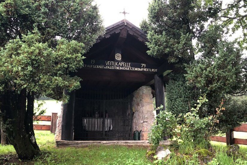 Veitlkapelle Eine kleine Kapelle, umgeben von grünen Bäumen und Pflanzen. Der Eingang ist mit einem Gitter versehen und ein Kreuz befindet sich auf dem Dach.