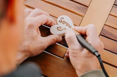 Eine Person graviert mit einem Werkzeug einen Schriftzug auf ein Herz aus Holz. Der Arbeitsplatz besteht aus einem Holztablett.