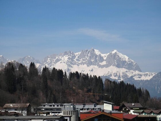 Aussicht auf das Bergmassiv Blick über Hausdächer und einen Nadelwald auf ein schneebedecktes Felsmassiv unter blauem Himmel.