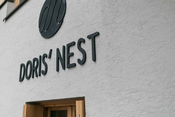 Fassade Doris' Nest Schwarzer Schriftzug Doris' Nest an einer hellen Hausfassade über einem Holztürrahmen.
