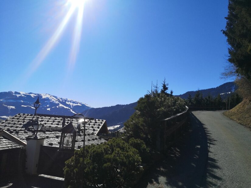 Asphaltierter Bergweg neben einem Hausdach mit Blick auf schneebedeckte Gipfel unter blauem Himmel.