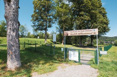 Eingangstor zu einer eingezäunten Hundewiese mit Willkommensschild und Bäumen im Hintergrund.