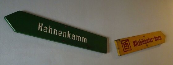 Zwei hölzerne Wegweiser an einer weißen Wand zeigen zum Hahnenkamm und Kitzbüheler Horn.