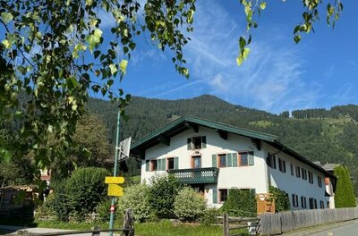 IMG_8763 Ein charmantes Haus umgeben von Bäumen und grüner Natur. Im Hintergrund sind sanfte Berge und ein klarer blauer Himmel zu sehen.