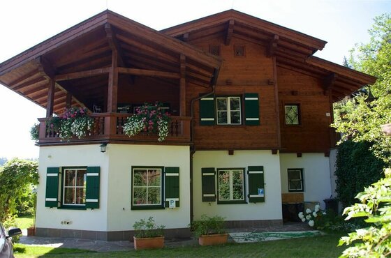 Zweistöckiges Alpenhaus mit weißer Fassade im Erdgeschoss, dunkler Holzverkleidung oben und grünen Fensterläden.