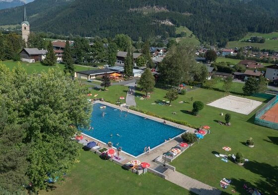 Ein schöner Pool in einer ländlichen Umgebung, umgeben von grünen Wiesen und Bergen. Im Wasser sind Menschen beim Schwimmen, und es gibt Liegen mit Sonnenschirmen an den Rändern.