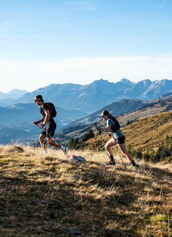 Zwei Trailrunner erklimmen einen hügeligen Pfad, umgeben von majestätischen Bergen und einer weiten, grünen Landschaft.