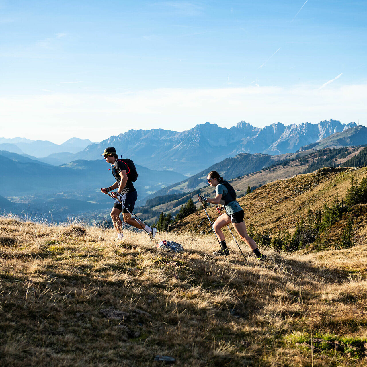 Sport Trailrunning Südberge Aurach (c) Kitzbühel Tourismus Zwei Trailrunner erklimmen einen hügeligen Pfad, umgeben von majestätischen Bergen und einer weiten, grünen Landschaft.