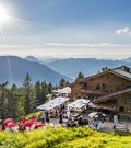 Berghütte aus Holz mit belebter Terrasse und Sonnenschirmen vor Gebirgspanorama bei Sonnenschein.