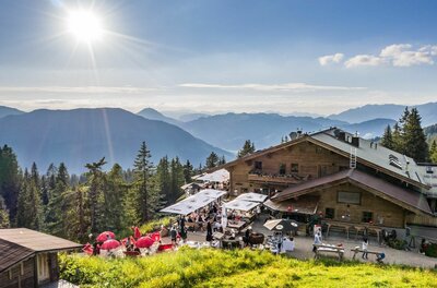 Berghütte aus Holz mit belebter Terrasse und Sonnenschirmen vor Gebirgspanorama bei Sonnenschein.