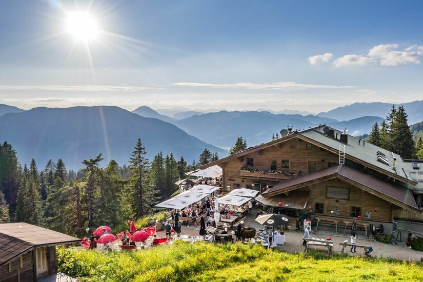 Hochbrunn Alm im Sommer Berghütte aus Holz mit belebter Terrasse und Sonnenschirmen vor Gebirgspanorama bei Sonnenschein.