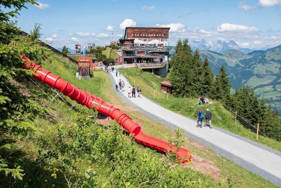 Berggasthof mit Erlebnisspielplatz Rote Röhrenrutsche an einem Hang neben einem Wanderweg und einem Berggasthof.